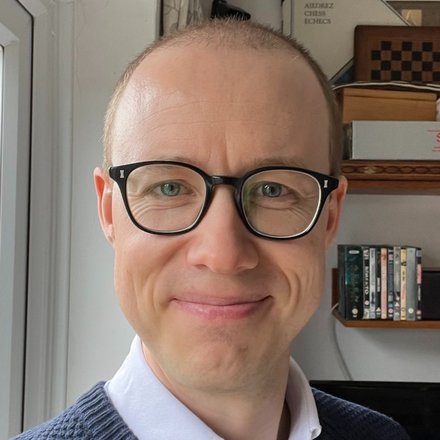 Chris Scrace smiling at the camera, wearing glasses and a navy jumper over a white collared shirt